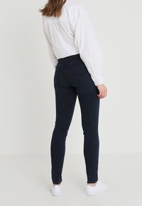 Jeans ajustados de denim azul oscuro, con una cintura alta, bolsillos traseros y una textura suave, combinados con una camisa blanca de botones y zapatillas blancas.