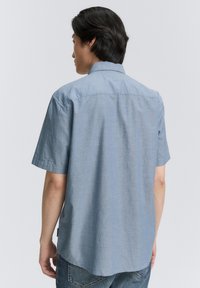 Cămașă albastră din chambray cu mânecă scurtă, guler cu buton, textură netedă și tiv curbat, asortată cu blugi din denim deschis.