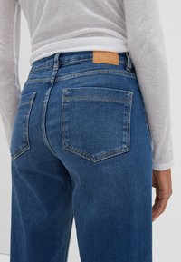 Jeans en denim bleu moyen avec une taille ajustée, des poches arrière et une étiquette en cuir à l'arrière. La texture semble lisse avec un minimum de décoloration.