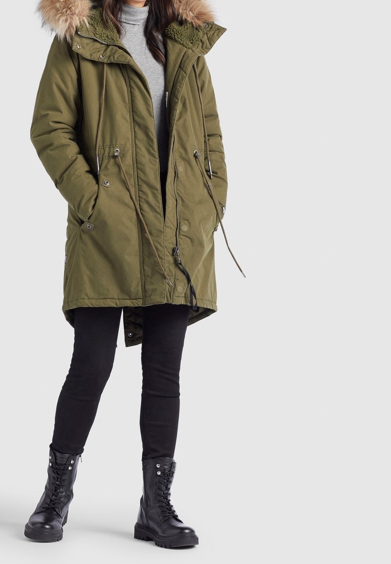 Personne portant une parka vert olive avec capuche en fourrure, un col roulé gris, un pantalon skinny noir et des bottes de combat noires à lacets, debout.