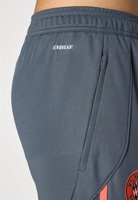 Graue Sportshorts aus strukturiertem Material, mit elastischem Bund, seitlichem Reißverschlusstaschen und einem Logo mit roten Akzenten.
