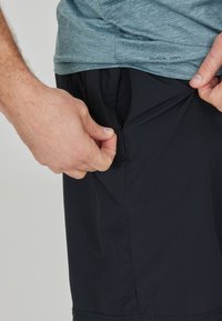 Schnell trocknende Sportshorts in Schwarz, mit einer seitlichen Reißverschlusstasche. Glatte Textur, leichtes Material, geeignet für aktive Verwendung.