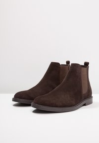 Botas Chelsea de ante marrón con paneles laterales elásticos, punta redondeada y suela de goma plana. Presentan una lengüeta para facilitar el calzado.