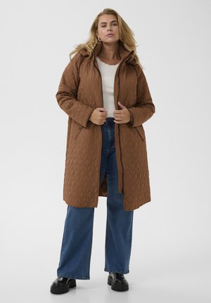 Femme portant un long manteau matelassé marron, un haut blanc, un jean bleu à jambes larges et des bottes noires épaisses sur un fond uni.