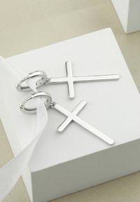 Silverkorsörhängen med en polerad yta, som har cirkulära hoop-fästen. Positionerade på en vit presentationslåda med ett band.