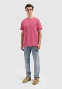 Rosa Baumwoll-T-Shirt mit kurzen Ärmeln und Rundhalsausschnitt; kombiniert mit hellblauen, verwaschenen Jeans und beigen Freizeitschuhen.