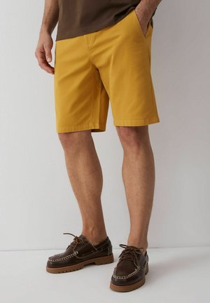 Mężczyzna ubrany w szorty do kolan w kolorze musztardowym, brązowe mokasyny na sznurowadła i brązową koszulę z jedną ręką w kieszeni.