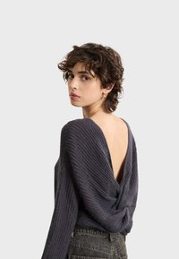 Gestrickter Pullover in Dunkelgrau mit drapiertem Rücken und tiefem V-Ausschnitt. Strukturiertes Material und lockere Passform, kombiniert mit hochtaillierten Jeans.