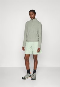Light green zip-up fleece top with a high collar, paired with matching shorts featuring cargo pockets. Model wears gray sneakers and dark socks.