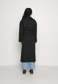 Manteau long noir avec une taille ceinturée, des manches longues et des détails à boutons, associé à un jean large bleu clair et des chaussures noires.