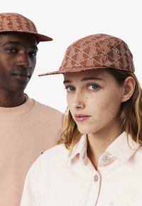 Cappello marrone a fantasia con visiera piatta. Due occhielli visibili, indossato da un modello con una maglietta rosa. Sfondo bianco semplice.
