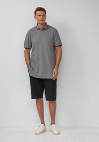 Grijze polo met zwarte accenten, gecombineerd met donkere shorts. Het model staat in witte sneakers en toont een casual, ontspannen uitstraling.