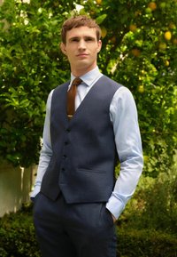 Dunkelblauer Weste über einem hellblauen Hemd mit strukturiertem Finish. Braune Krawatte mit Punktenmuster. Taschen an der Weste, taillierte Passform.