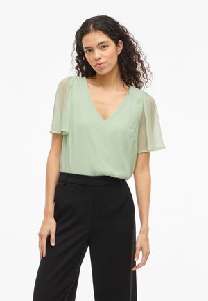 Woman with curly hair wears light green V-neck blouse with sheer short sleeves and high-waisted black trousers against white background.