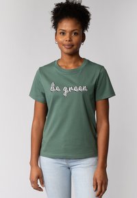 T-shirt en coton vert à manches courtes, affichant le texte "sois vert" en blanc, avec des vagues subtiles. Texture lisse et coupe classique.