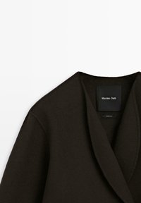 Ciemnobrązowy wełniany płaszcz z kołnierzem typu szal, o fakturowanym wykończeniu i metką po lewej stronie. Prosty, nowoczesny design.