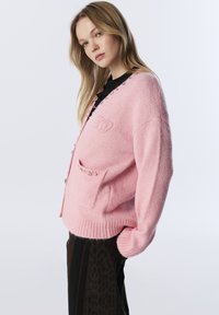 Różowy kardigan z mieszanki wełny z kieszenią z przodu, dekoltem w kształcie V oraz dekoracyjnymi guzikami wzdłuż zapięcia. Zawiera haftowane logo.
