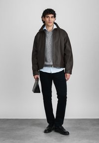 Jaqueta de couro castanho-escuro sobre um suéter cinza texturizado, combinada com calças pretas e sapatos pretos. O modelo segura uma bolsa pequena. Fundo simples.