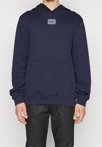 Sweat à capuche bleu marine avec une poche kangourou, un tissu texturé et un écusson logo rectangulaire de couleur contrastante sur la poitrine.