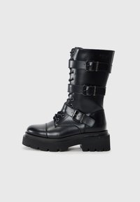 Black leather knee-high boot featuring lace-up front, multiple adjustable straps, thick platform sole, and rounded toe.