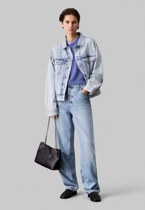Young person standing in light denim jacket and jeans, lavender sweater, black shoes, holding black handbag with chain straps.