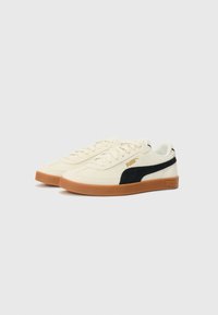 Ténis Puma com um upper de creme, faixa lateral preta e sola de borracha caramelo. Apresenta atacadores planos e um acabamento em tecido texturizado.