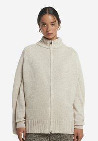 Krämfärgad cardigan i stickad kvalité med hög krage, hel dragkedja och ribbade muddar och nederkant. Har en lös, oversized passform.