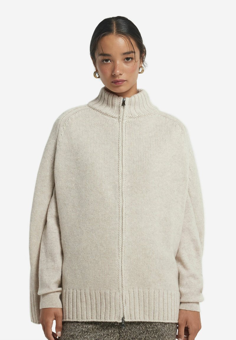 Krämfärgad cardigan i stickad kvalité med hög krage, hel dragkedja och ribbade muddar och nederkant. Har en lös, oversized passform.