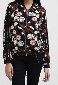 Sort lynlåsjacket med pink og hvide blomster- og bladmønstre, båret med sorte bukser.