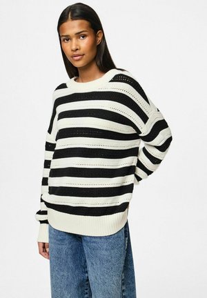 Svart och vit randig stickad tröja med chunky ribbad textur, vida ärmar och rundad halsringning. Avslappnad passform, bärs med blå jeans.