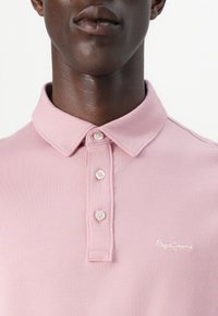 Πόλο μπλούζα σε ανοιχτό ροζ ύφασμα με κοντά μανίκια, γιακά και μπροστινή ταινία με τρία κουμπιά. Διαθέτει κεντημένο λογότυπο "Pepe Jeans" στο στήθος.