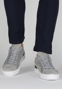 Zapatillas de ante gris con suelas de goma blanca, que presentan un diseño de cordones gris y acentos en el talón en negro. Los pantalones son de un azul oscuro, con corte ajustado.