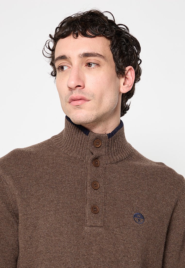 Maglione di lana marrone con colletto alto, tre bottoni marroni sul colletto e rivestimento interno blu navy. Presenta un piccolo logo sul petto.
