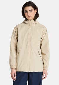 Beige lättviktsjacka med huva, elastiska ärmslut, framdragkedja och sidofickor, tillverkad av slät, vattenavvisande tyg.