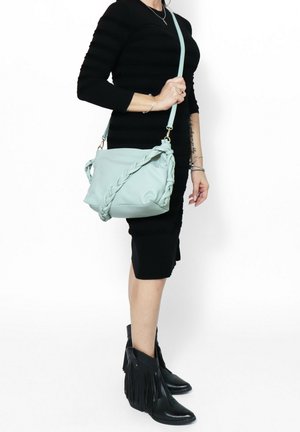 Hellblaue Leder-Crossbody-Tasche mit geflochtenen Details, goldenem Beschlag und verstellbarem Träger, kombiniert mit schwarzen Fransen-Stiefeletten.