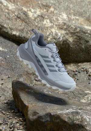 Gray and white Merrell trail running shoe hovering above rocky, uneven outdoor terrain with gravel and large stones.