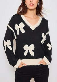 Pull en tricot noir avec col en V, motifs de nœuds blancs et bordure crème contrastante au niveau de l'encolure et de l'ourlet. Texture douce et coupe décontractée.