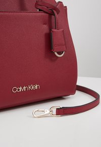 Sac à main rouge Calvin Klein avec logo doré, texture en cuir et bandoulière amovible avec boucle dorée sur fond blanc.
