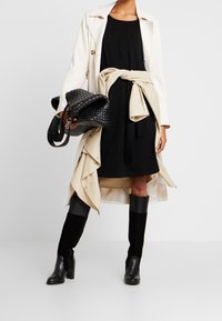 Femme portant une robe noire avec une veste beige nouée à la taille, un trench-coat blanc, des bottes noires montant jusqu'aux genoux et tenant un sac à main noir tissé.