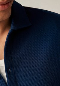 Chemise à manches courtes bleu marine avec col, en tissu texturé, présentant un détail de boutonnière visible et un sous-pull blanc contrastant en dessous.