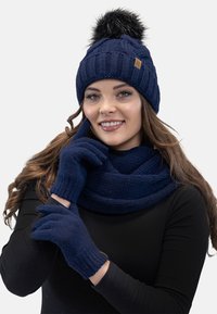Donkerblauwe gebreide beanie met een nep bont pom-pom, bijpassende handschoenen en een sjaal, allemaal met een textuurpatroon, weergegeven tegen een lichte achtergrond.