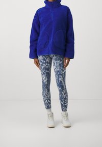 Kungablå fleece-pullover med zip-krage, oversized passform och två stora framfickor, matchad med mönstrade blå leggings och vita sneakers.