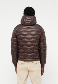 Braune Pufferjacke mit wellenförmigem Steppmuster, glänzender Textur und hohem Kragen, kombiniert mit cremefarbenen Hosen.