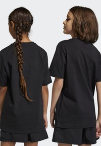 T-shirt in cotone nero con maniche corte, vestibilità rilassata e scollatura rotonda. La vista posteriore mostra un taglio dritto e una linea dell'orlo svasata. Indossato con pantaloni corti neri.