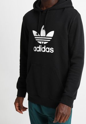 Černá mikina Adidas s bílým logem trefoil, kterou nosí osoba s tmavou pokožkou a tyrkysovými kalhotami na jednobarevném pozadí.