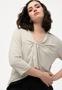Langærmet, lys beige bluse med en snoet knudedetalje foran og v-udskæring. Glat stof, afslappet pasform, parret med sorte bunde.