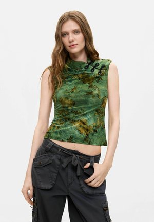 Vrouw draagt een groene mouwloze top met bloemenprint, zwarte decoratieve knopen en zwarte cargobroek met zakken en een trekkoordriem.
