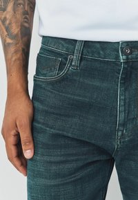 Tumedat türkiissinised teksapüksid kitsase lõikega. Detailide hulka kuuluvad esikäe nööpidega sulgur, kontrastne õmblus ja tagatasku, millel on logo detail.