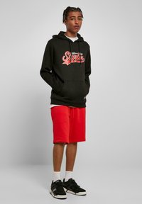 Sweatshirt à capuche noir avec un imprimé graphique rouge, associé à un short rouge. Le modèle porte des baskets noires et des chaussettes blanches montantes. Textures lisses.