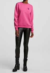 Sudadera rosa con cuello redondo y pequeño diseño bordado en el pecho, combinada con leggings negros brillantes y botas de tobillo negras gruesas.
